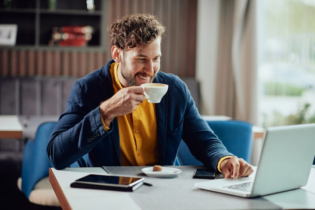 Close up of smiling freelancer sitting in cafe, drinking fresh coffee and using laptop