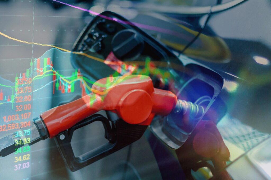 red fuel nozzle inserted into car's fuel tank; stock market graphs and numbers; conveying themes of fuel prices and economic impact